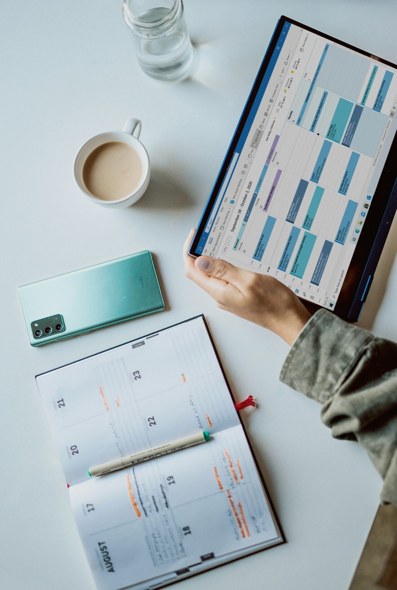 Hand houdt een tablet vast met een digitale agenda, naast een papieren planner, een telefoon en een kopje koffie op een bureau