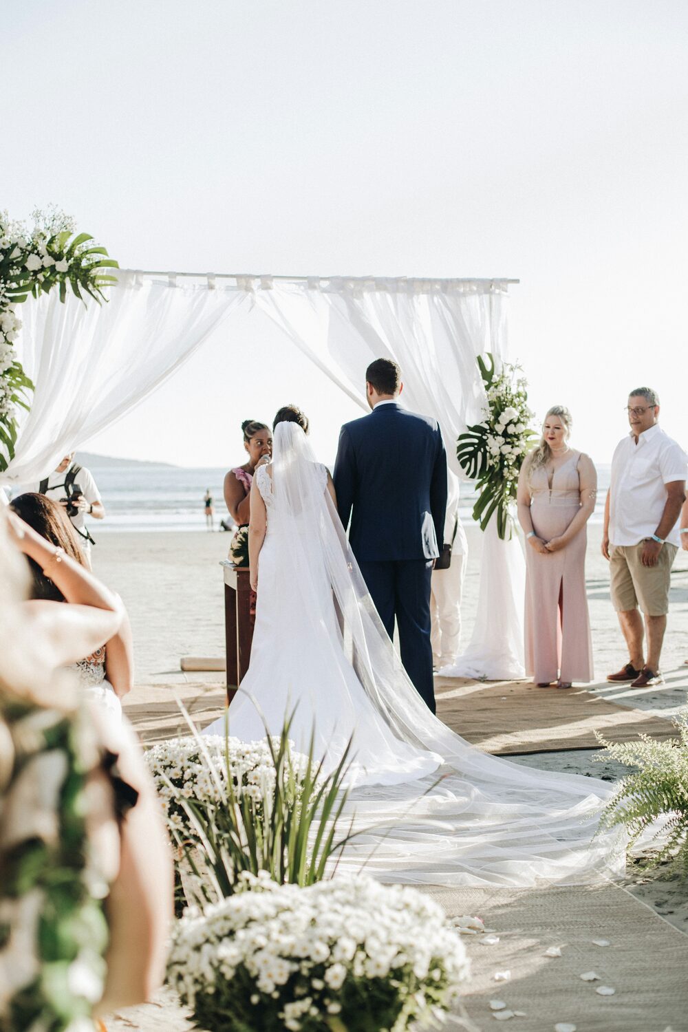 Bruidspaar tijdens een strandceremonie onder een witte boog met bloemen