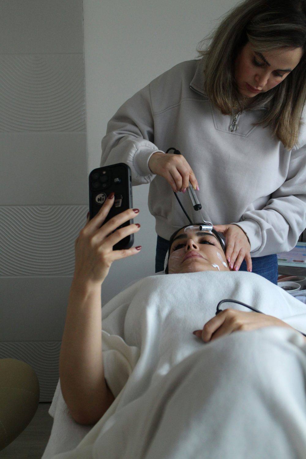 Klant kijkt op haar telefoon tijdens een gezichtsbehandeling in de salon