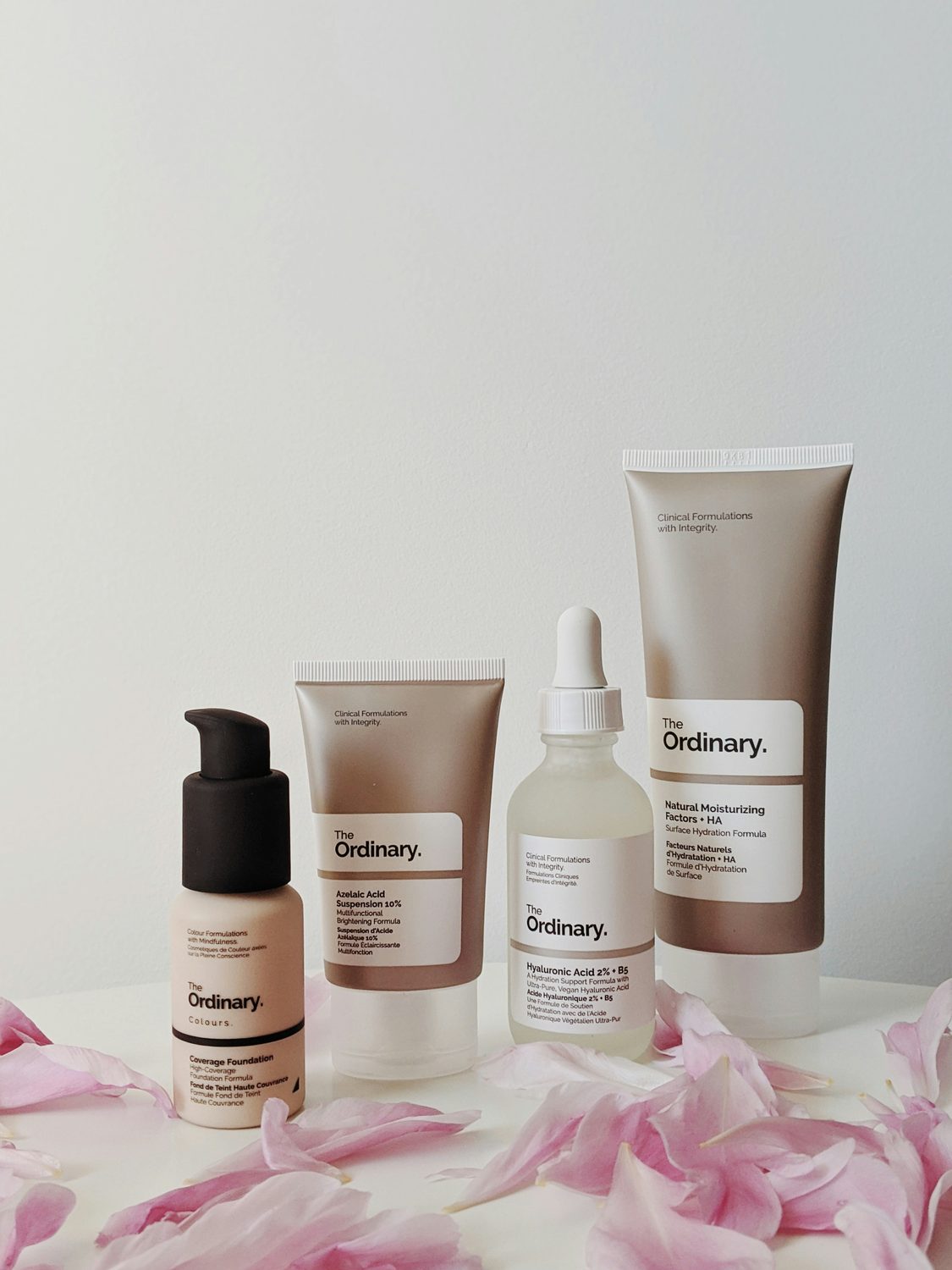 Skincare producten voor een reisroutine op een rij met roze bloemblaadjes, waaronder een serum en een hydraterende creme