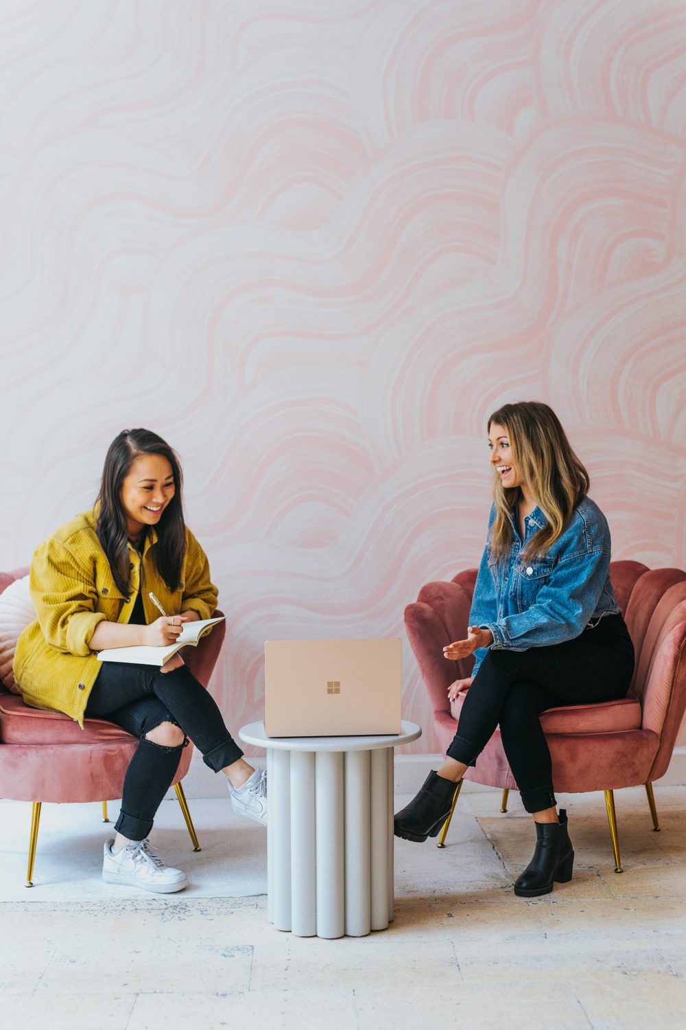 Twee beauty-ondernemers overleggen lachend in roze fauteuils met een laptop tussen hen in